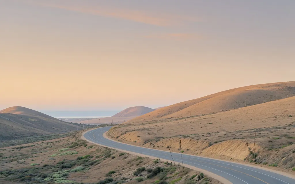 A quiet open coastal road curving through San Diego County hills toward soft morning light